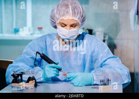 Un technicien dentaire en vêtements de protection travaille sur une dent prothétique dans son laboratoire. Banque D'Images