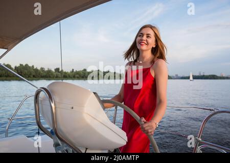 Capitaine de femme de pritty dans une longue robe rouge tenant la barre du navire. Fille de navigation d'un yacht Banque D'Images