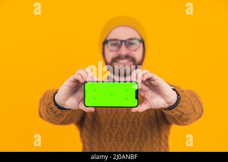 Portrait d'un jeune homme caucasien souriant barbu montrant l'écran vert horizontal de son smartphone à l'appareil photo, tenant le téléphone avec deux mains sélective focus est sur le téléphone et les mains décontracté attire orange arrière-plan isolé studio tourné . Photo de haute qualité Banque D'Images