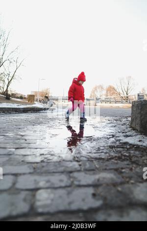 bonne fille de bébé en bottes et veste rouge courir et jouer dans la flaque d'eau. drôle gai fille de tout-petit jouer à l'extérieur Banque D'Images