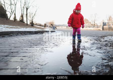 bonne fille de bébé en bottes et veste rouge courir et jouer dans la flaque d'eau. drôle gai fille de tout-petit jouer à l'extérieur Banque D'Images