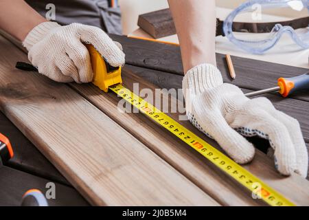 Le menuisier mesure la longueur des planches de bois ou des poutres en bois en mesurant le ruban ou la règle. Menuisier espace de travail, artisan entrepreneur. Mains dans des gants de JO Banque D'Images