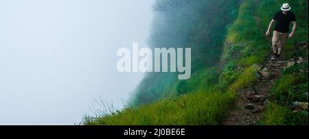 Homme blanc avec chemise noire et chapeau marchant le long d'une profonde falaise brumeuse au-dessus des nuages sur la montagne au cap-vert Banque D'Images