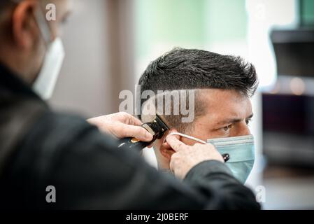 Crop barber dans le masque avec le coupe-cheveux faisant une coupe de cheveux à un client masculin dans le salon de coiffure pendant l'épidémie de COVID 19 Banque D'Images