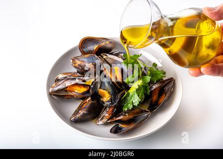 Moules cuites avec feuilles de persil dans un plat blanc recouvert d'huile d'olive extra vierge versée à partir de carafe en verre sur fond blanc Banque D'Images