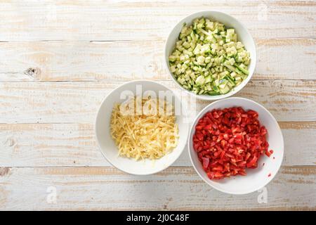 Légumes en dés, courgettes et poivron rouge, fromage râpé dans des bols blancs sur une table rustique en bois, espace copie, vue d'en haut, sel Banque D'Images