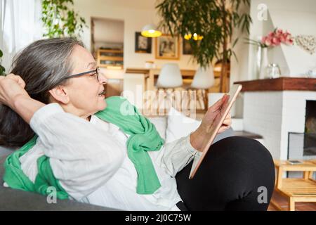 Une vieille femme sur le canapé à la maison ou à la retraite, discutant vidéo sur la tablette Banque D'Images