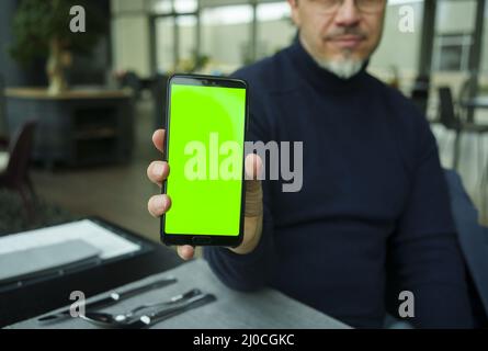 Homme assis dans un restaurant ou un café en utilisant le téléphone avec écran vert vierge. Restaurant, application de commande de nourriture. Banque D'Images