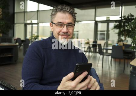 Homme souriant d'âge moyen utilisant le téléphone assis dans le restaurant ou le café. Banque D'Images