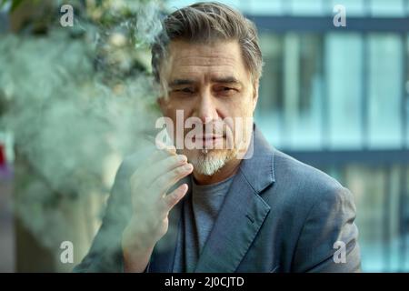 Homme d'âge moyen fumant un cigare à l'extérieur devant le bâtiment de bureau, pensant, regardant loin. Banque D'Images