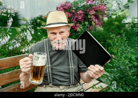 un vieil homme aux cheveux gris dans un chapeau avec un ordinateur portable boit de la bière. Banque D'Images