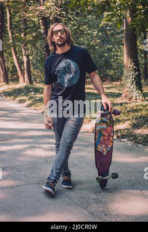Longboarder se posant à côté de son long skateboard sur la route dans la forêt. Patinage à planche Banque D'Images