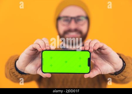 Jeune homme caucasien gai barbu montrant l'écran vert horizontal de son smartphone à l'appareil photo, tenant le téléphone avec deux mains sélective focus est sur le téléphone et les mains décontracté attire orange arrière-plan isolé studio tourné de gros plan . Photo de haute qualité Banque D'Images