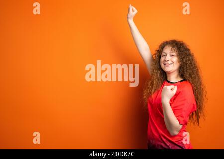 Fan football redhead femme dans le t-shirt rouge uniforme gardant le poing lever la main et une autre main se mettre au coude. Banque D'Images
