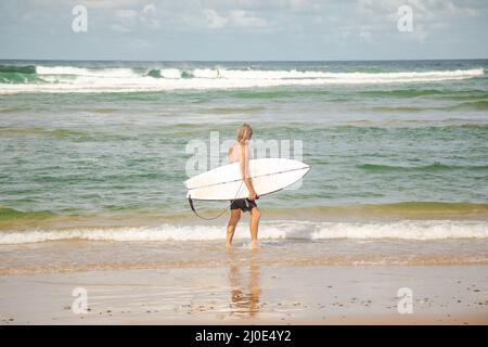 Un homme de surf âgé aux cheveux longs marchant le long du bord de l'eau transportant des planches de surf à Coffs Harbour, Nouvelle-Galles du Sud, Australie Banque D'Images