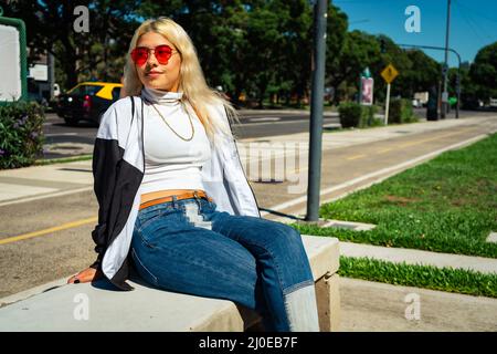 Belle jeune femme latine dans un t-shirt blanc assis sur un banc dans un parc public. Concept de femme moderne. Banque D'Images