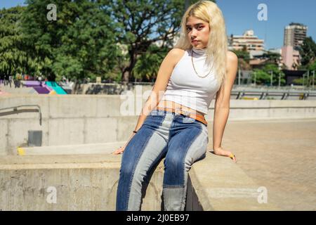 Une belle jeune femme Latina dans un t-shirt blanc assis sur une rampe de ciment tout en regardant sérieusement en avant sur une journée ensoleillée. Banque D'Images