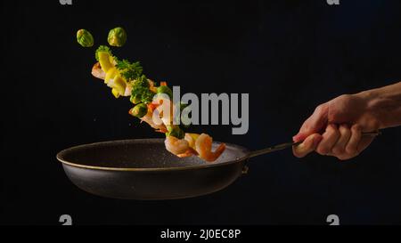 Crevettes tigrées avec des tranches de citron, des légumes et des herbes dans une casserole dans un vol gelé sur fond noir. Processus de cuisson. Cuisine asiatique, cuisine gastronomique. Banque D'Images
