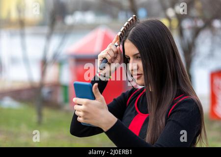 Adorable jeune fille se brossant les cheveux et souriant agréablement tout en regardant le smartphone comme un miroir dans un parc Banque D'Images