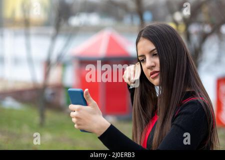 Adorable jeune fille se brossant les cheveux et souriant agréablement tout en regardant le smartphone comme un miroir dans un parc Banque D'Images