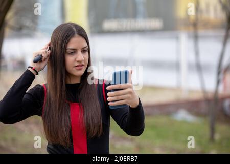 Adorable jeune fille se brossant les cheveux et souriant agréablement tout en regardant le smartphone comme un miroir dans un parc Banque D'Images