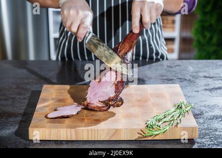 Un cuisinier coupe un agneau fumé en tranches sur une planche à découper en bois Banque D'Images