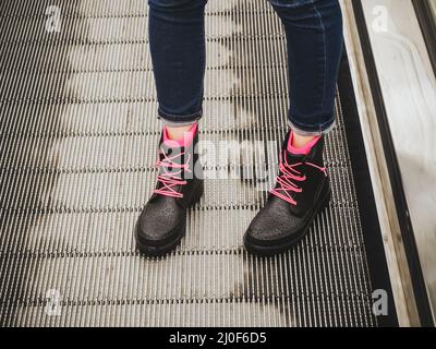 Les jambes des femmes en Jean bleu et les bottes noires en caoutchouc humide avec lacets roses se tiennent sur l'escalier roulant Banque D'Images