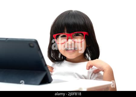 Happy Little Asian Preschooler fille portant des lunettes rouges et utilisant le PC de tablette sur fond blanc et sourire, asiatique fille apprenant avec un appel vidéo avec Banque D'Images