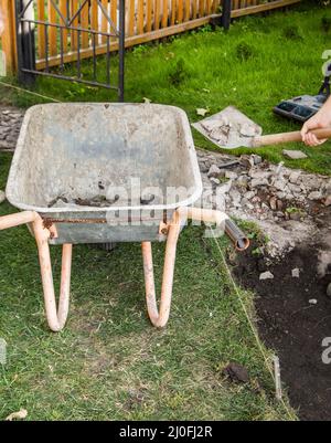 Une vieille brouette rouillée vide pour l'enlèvement de la terre pendant la construction d'un nouveau chemin de jardin, à l'ouverture, en été, arrière-cour. Banque D'Images