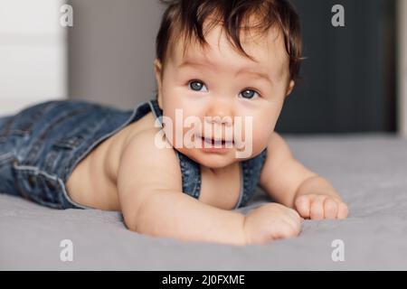 Portrait d'un bébé portant une barboteuse en denim couché sur une couverture grise à la maison. En regardant les yeux bleus de bébé enfant ramper sur le lit dans la chambre, explorez le monde Banque D'Images