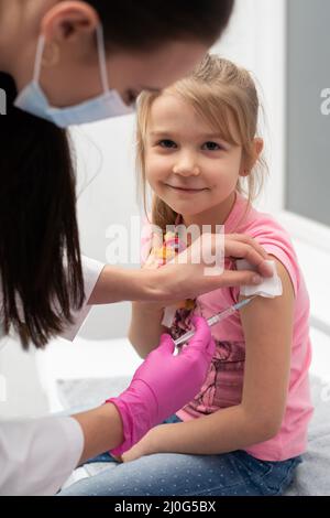 Tout en recevant le vaccin, la jeune fille souriante regarde directement dans la lentille. Le médecin appuie l'enfant dans le bras avec une aiguille. Banque D'Images