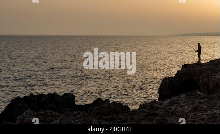 Silhouette d'un pêcheur pêcheur pêchant dans l'océan à coucher de soleil Banque D'Images