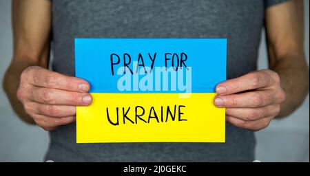 Un homme debout sur un gris, tient dans ses mains le fond d'un drapeau ukrainien en carton jaune-bleu avec l'inscription 'prier pour l'Ukraine' Banque D'Images