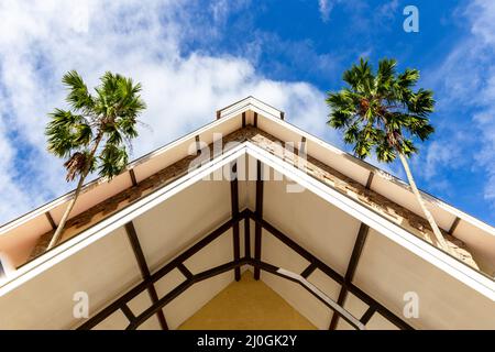 Toit d'église de style colonial, vue symétrique à angle bas avec deux palmiers à noix de coco sur les côtés et ciel bleu en arrière-plan Banque D'Images