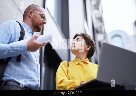 Vue de dessous des jeunes collègues à l'extérieur discutant à l'extérieur de l'immeuble de bureaux avec plans et ordinateur portable. Équipe d'affaires de designers, gestionnaires immobiliers dans des vêtements décontractés brainstorming à l'extérieur Banque D'Images