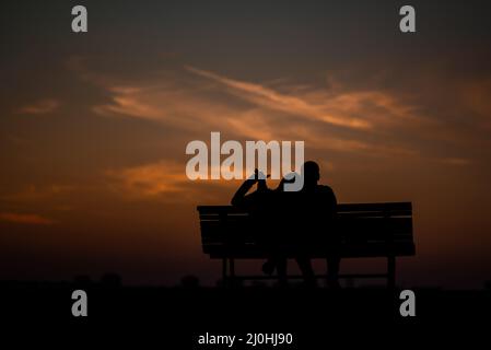 Silhouettes d'un couple assis sur une chaise contre un ciel de soirée coloré Banque D'Images