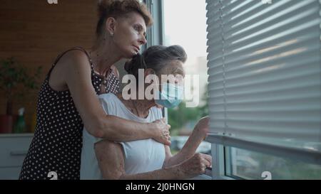 Femme solitaire âgée de 90 ans dans un masque médical et sa fille mature debout dans embrasse regardant la fenêtre dans la maison de soins infirmiers. Banque D'Images