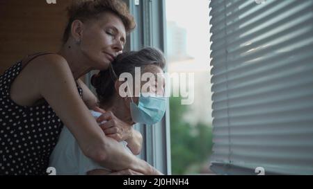 Fille adulte embrassant avec la mère aînée aimante 90 ans dans le masque debout à la fenêtre, des parents de différentes générations, WO Banque D'Images