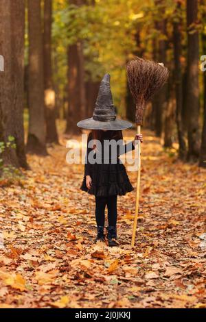 Jolie petite fille à cheveux rouges vêtue d'un costume de sorcière debout avec un balai sur fond de forêt d'automne Banque D'Images