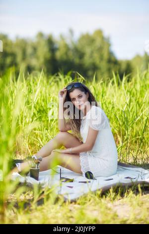 Jeune femme blonde en robe blanche et lunettes de soleil est assise sur une couverture en herbe haute. Banque D'Images