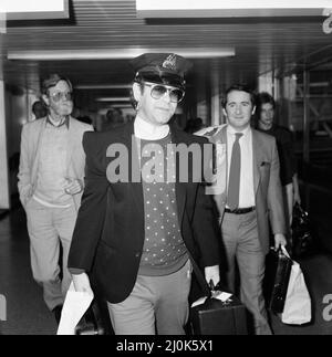 Le chanteur Elton John arrive à l'aéroport d'Heathrow après sa visite des États-Unis d'une durée de 10 semaines, portant un chapeau de policier américain. Sur la photo à droite se trouve son directeur John Reid. 21st août 1982. Banque D'Images