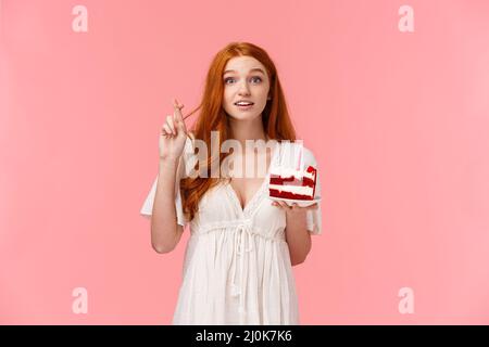 Mignonne et tendre, jolie fille à tête rouge en robe blanche, célébrant l'anniversaire, signe de la croix bonne chance, tenant le gâteau du jour Banque D'Images