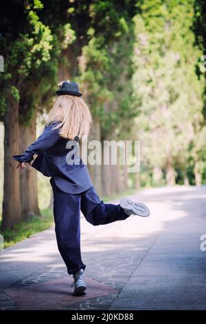 Femme vêtue de bleu et chapeau dans une posture dansante sur une allée du parc. Danseur de rue Banque D'Images