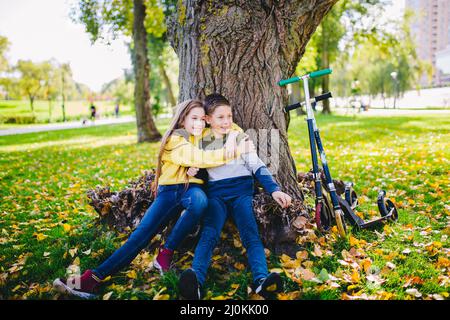 Un garçon et une fille de race blanche s'embrasent sous un arbre dans le parc après avoir fait du scooter en automne. Portrait de frère et sis Banque D'Images