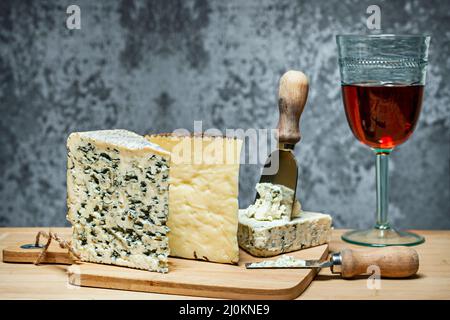 Le fromage reste encore en vie avec brie, coin manchego et bleu salé français sur une planche en bois avec des couteaux à fromage et un verre de vin complet Banque D'Images