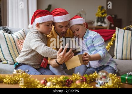 Un père caucasien et deux fils ouvrent une boîte cadeau pendant un appel vidéo sur un smartphone pendant noël Banque D'Images