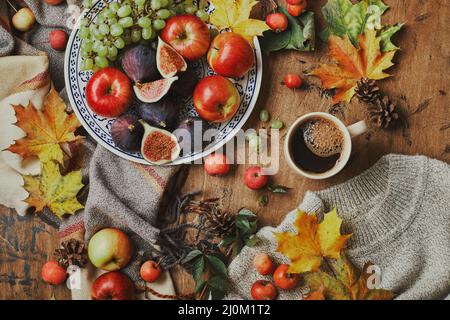 Plat avec figues, pommes et raisins et tasse de café sur fond de bois avec un pull chaud, écharpe, feuilles d'automne et pommes. A Banque D'Images