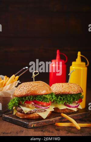 Deux hamburgers faits maison avec du bœuf, du fromage et de la marmelade d'oignons sur une planche de bois, des frites dans un panier métallique et des sauces. Conc. Restauration rapide Banque D'Images