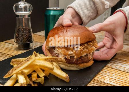 en tenant deux mains un délicieux hamburger de bœuf avec beaucoup de fromage à la crème et des oignons caramélisés à côté d'une pile de frites maison, un peu de poivre et un froid Banque D'Images