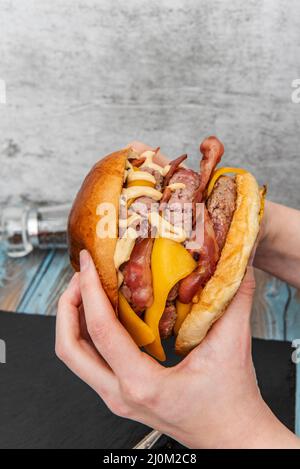 tenant entre deux mains un hamburger de bœuf à trois viandes avec beaucoup de bacon frit et du fromage cheddar accompagné de sauces Banque D'Images